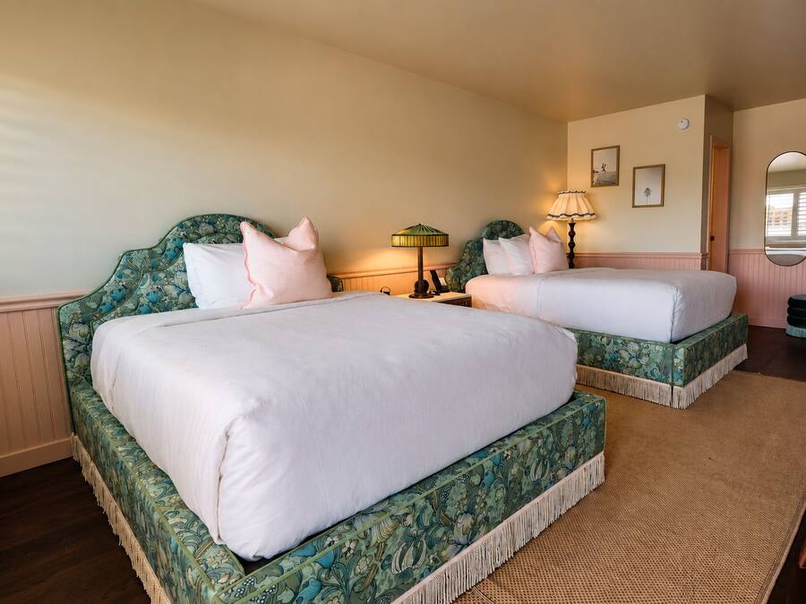 A hotel room with two beds, a nightstand, a lamp, a mirror, and a small table.