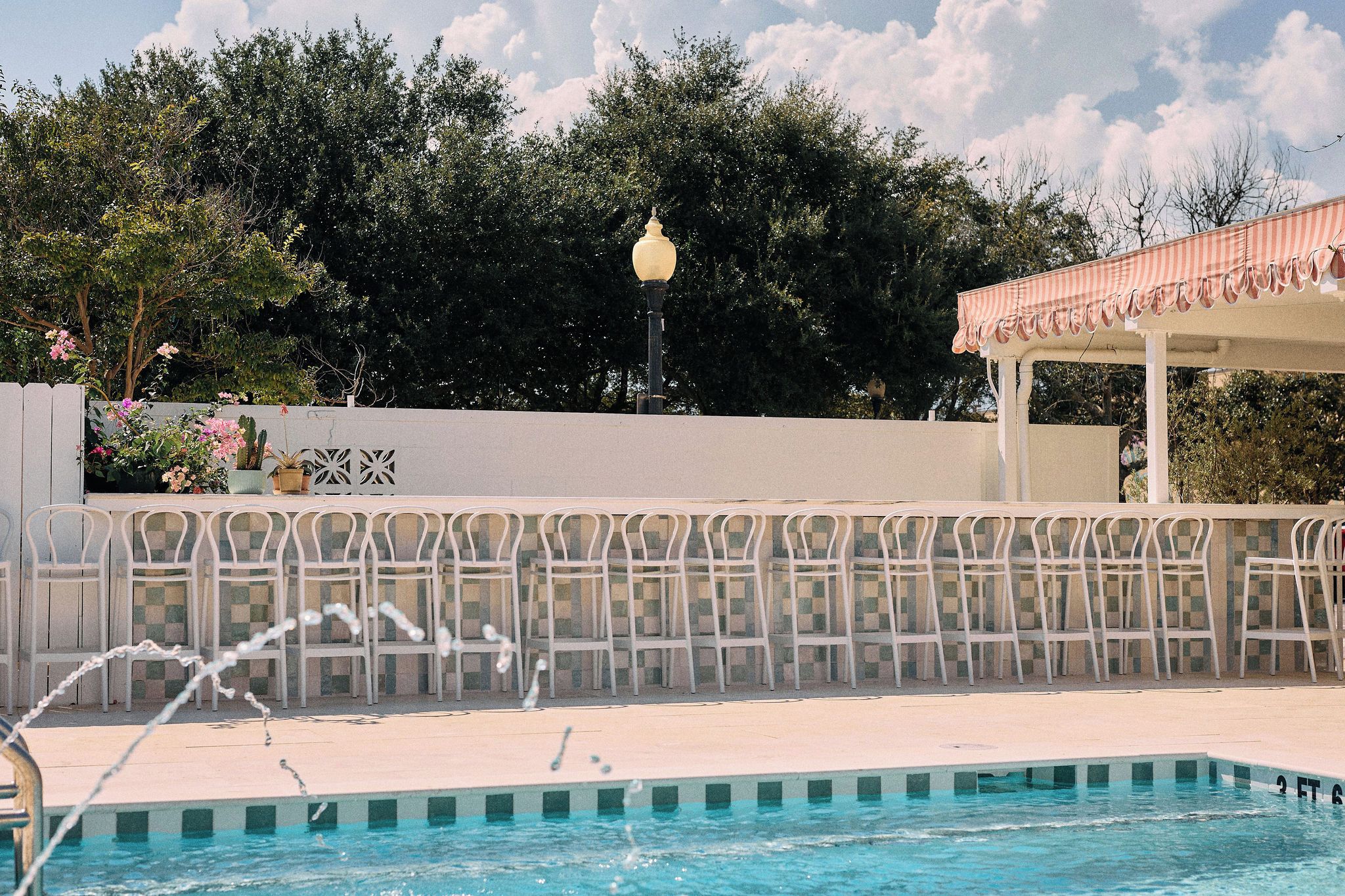 Ein Swimmingpool mit einer Fontäne, umgeben von einem weißen Zaun und einer Pergola mit gestreiftem Sonnensegel.