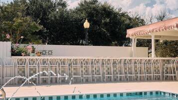 Ein Swimmingpool mit einer Fontäne, umgeben von einem weißen Zaun und einer Pergola mit gestreiftem Sonnensegel.