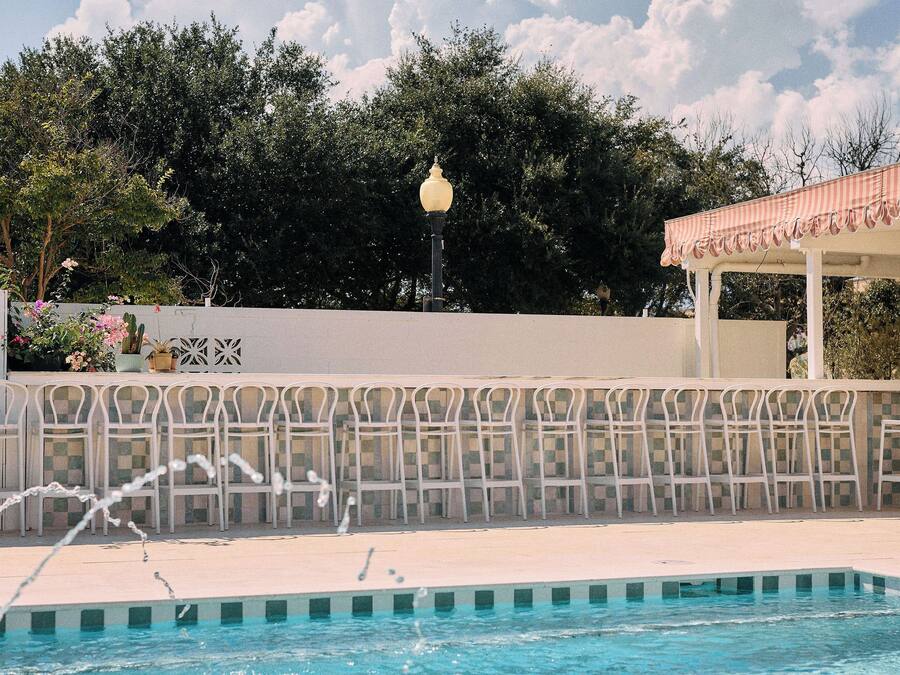 Ein Swimmingpool mit einer Fontäne, umgeben von einem weißen Zaun und einer Pergola mit gestreiftem Sonnensegel.