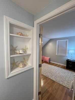 Ein Zimmer mit einer eingebauten Regalanlage, die Muscheln und Seesterne zeigt, einem gemusterten Teppich, einem Bett mit Kopfteil, einem Nachttisch mit Lampe und einem Fenster mit Jalousien.