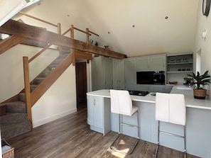 A kitchen with a wooden staircase, grey cabinets, a white countertop, and a small island with bar stools. - Ash Tree Lodge (Norwich)