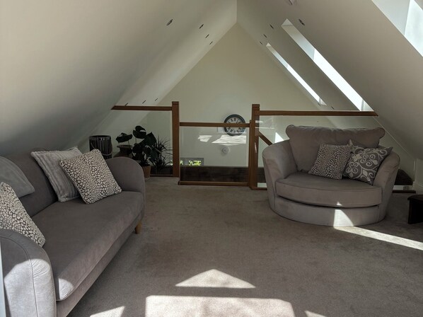A loft-style room with a sloped ceiling, two sofas, a wooden-framed window, and a potted plant.