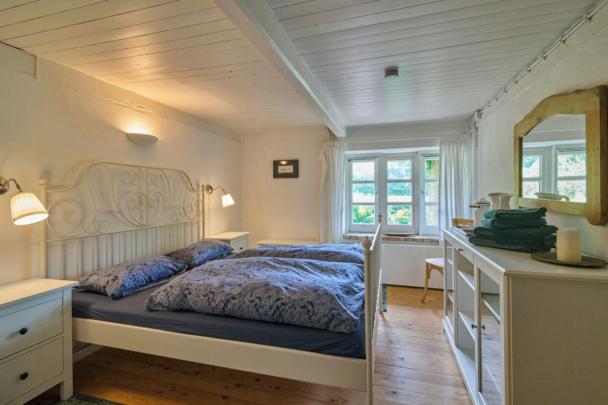 A bedroom with a bed, bedside lamps, a dresser, a window with curtains, and a mirror.