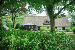 A rustic thatched-roof house with a covered outdoor seating area, surrounded by lush greenery. - Charming country house in Schleswig-Holstein (Fedderingen)