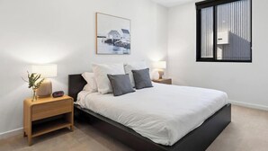 Ein modernes Schlafzimmer mit einem großen Bett, Nachttischen mit Lampen, einem gerahmten Bild an der Wand und einem Fenster mit Jalousien.
