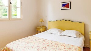 Un dormitorio con cama, mesita de noche, lámpara, un cuadro enmarcado y una ventana.