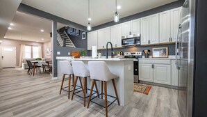 A modern kitchen with white cabinets, a stainless steel refrigerator, and a bar area with stools. - Luxurious, Modern Duplex | 6 Bedrooms! Sleeps 12+ (Indianapolis)
