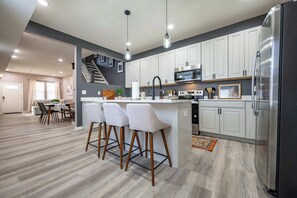 A modern kitchen with white cabinets, a stainless steel refrigerator, and a bar area with stools. - Luxurious, Modern Duplex | 6 Bedrooms! Sleeps 12+ (Indianapolis)