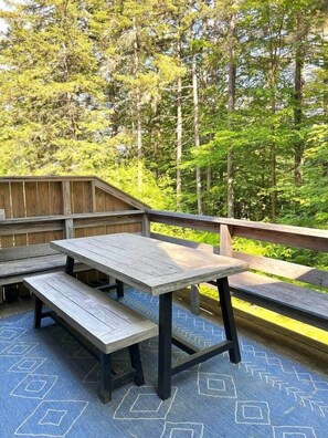 Eine Holzterrasse mit Picknicktisch und Bänken, umgeben von Bäumen.