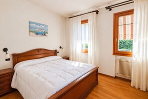 A bedroom with a wooden bed, a nightstand, a wall-mounted lamp, a window with curtains, and a radiator. - Between Prosecco Hills and Venice-Casa Il Sogno (Cal Grande)