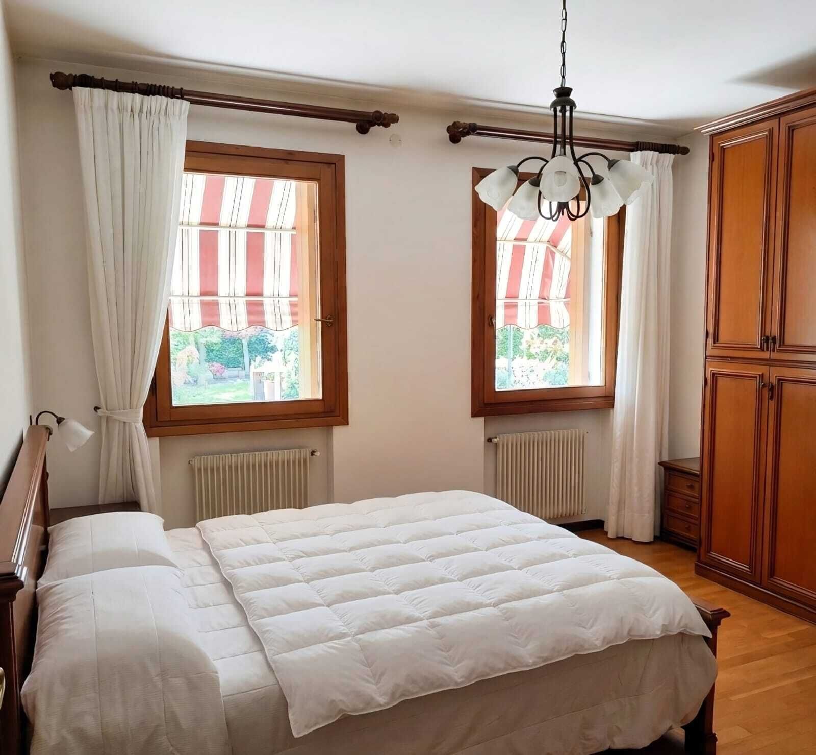 Une chambre à coucher avec un lit, deux fenêtres avec des rideaux, un radiateur et une armoire.