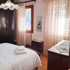 Quarto com cama, mesa de cabeceira, janela com cortinas, radiador e planta em vaso.