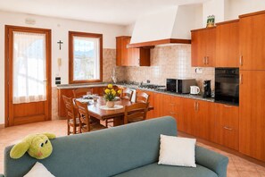 A kitchen with wooden cabinets, a dining table with chairs, a sofa, and a window with curtains. - Between Prosecco Hills and Venice-Casa Il Sogno (Cal Grande)