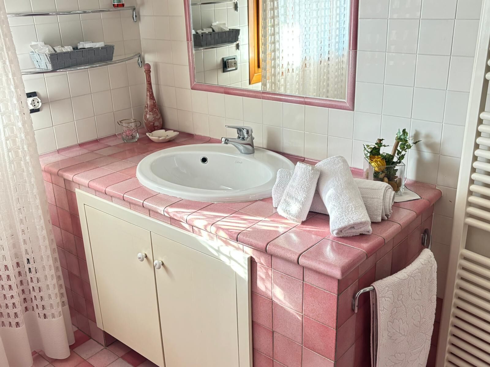 Un baño con tocador de azulejos rosas, un lavamanos blanco, un espejo y un perchero para toallas.