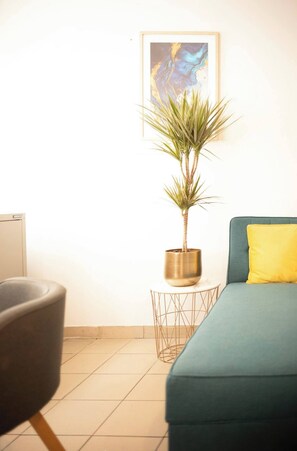 A room with a teal sofa, a yellow pillow, a plant in a metallic pot, and a framed abstract painting on the wall.