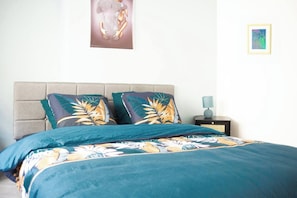 A bed with a floral bedspread, two pillows with tropical leaf patterns, a bedside lamp, and a framed artwork on the wall.