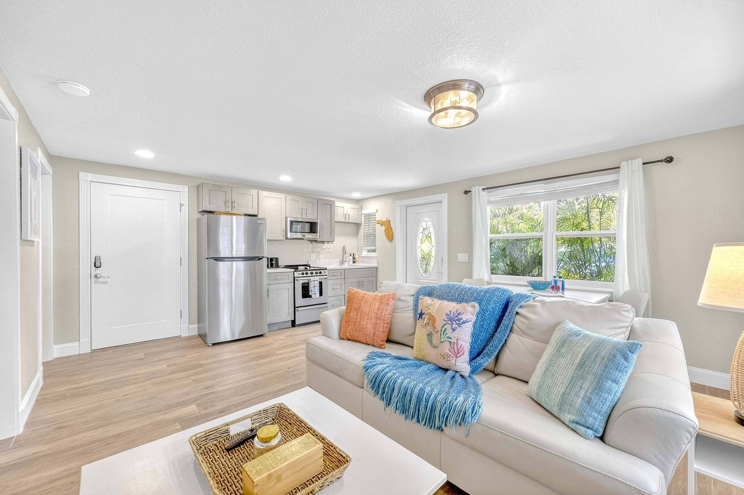 A modern kitchen with stainless steel appliances, a dining area with a white table and a basket, and a living room with a beige sofa and colorful cushions.
