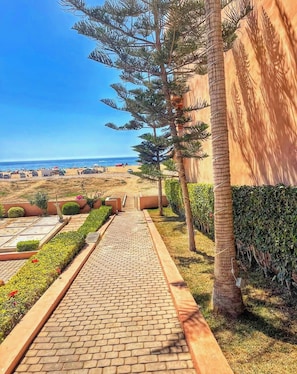 A paved pathway leading to a beach with a clear view of the ocean, surrounded by greenery and palm trees. - Dar Yanis -(2-4 Guests) -Beach on foot - A/C - Terrace - Parking - 2 Netflix TVs (Imi Ouaddar)