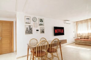 A living room with a wooden dining table, chairs, a sofa, and a TV displaying 'NETFLIX'. - Dar Yanis -(2-4 Guests) -Beach on foot - A/C - Terrace - Parking - 2 Netflix TVs (Imi Ouaddar)