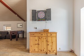 A room with a dartboard mounted on the wall, a wooden cabinet below, and a foosball table in the background.