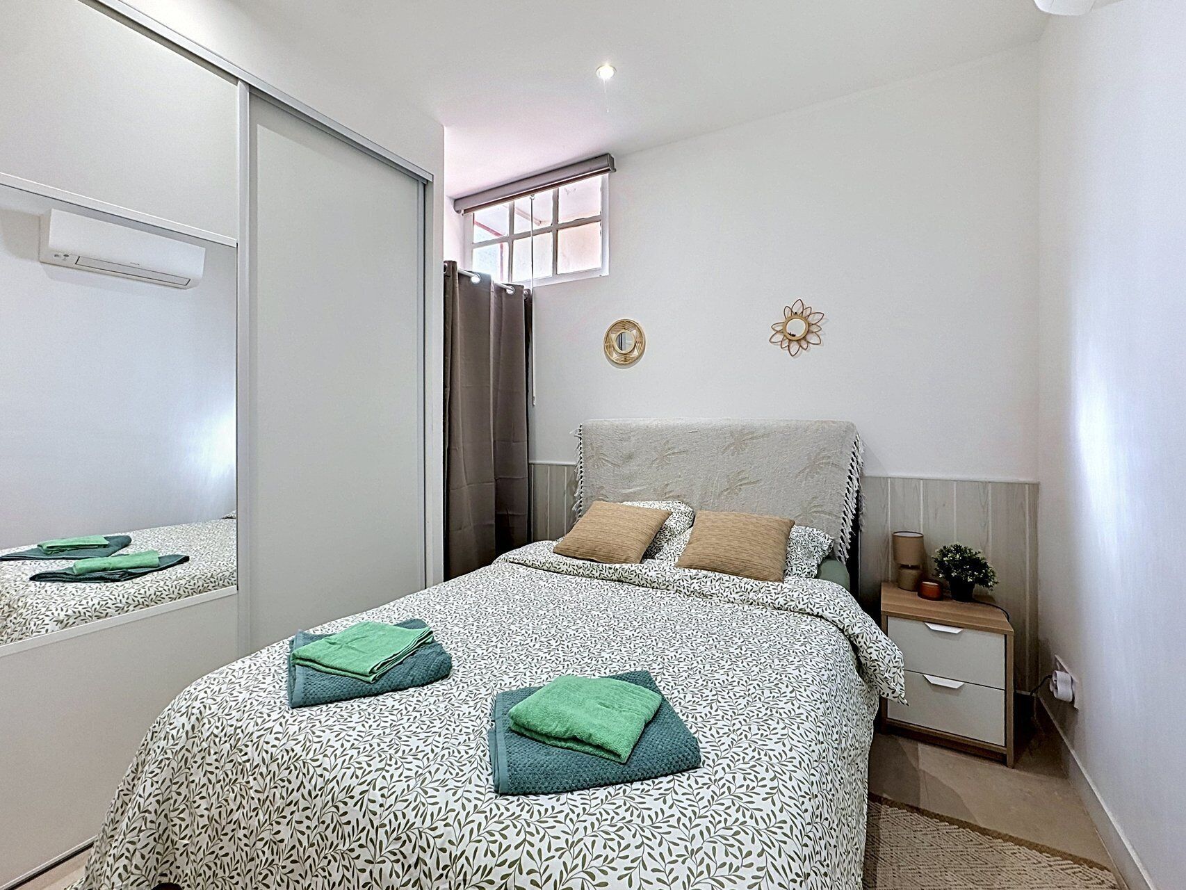 Een slaapkamer met een bed, nachtkastje, spiegel, raam met gordijnen en een wandklok.