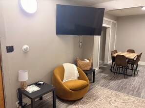A living room with a sofa, a table, a TV mounted on the wall, and a dining area with chairs.