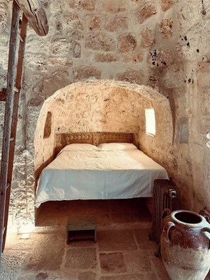 Ein Zimmer mit Steinwänden, einem Einzelbett, einem kleinen Tisch und einer großen Vase.