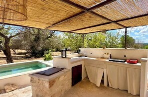 Eine überdachte Außenküche mit Pool, einer Steinarbeitsplatte und Blick auf Bäume.