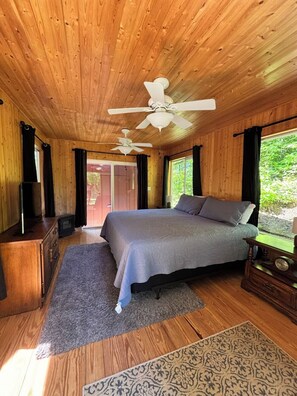 Ein Schlafzimmer mit Holzvertäfelung, einem Bett, zwei Deckenventilatoren, einem Fernseher und einem Fenster mit Vorhängen.
