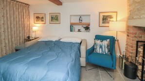 A bedroom with a bed, a chair, a fireplace, and wall-mounted shelves with decorative items. - The Cottage (Swindon)
