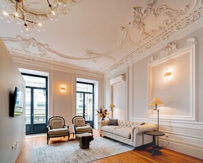 Ein geräumiges Wohnzimmer mit hoher Decke, aufwendigen Deckenverzierungen, einem großen Fenster und einem an der Wand montierten Flachbildfernseher.