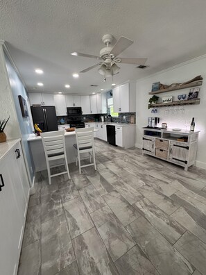 Eine moderne Küche mit weißen Schränken, einem schwarzen Kühlschrank, einem Essbereich mit zwei weißen Stühlen, einem Deckenventilator und einem Weinkühlschrank an der Wand.