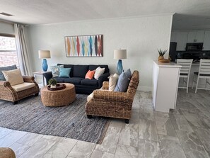 Ein Wohnzimmer mit einer dunklen Couch, zwei Korbstühlen, einem Couchtisch und einer Küche im Hintergrund.