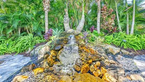 A garden with a small waterfall, palm trees, and various tropical plants. - Walk to Siesta Key Beach & Village | Pool | Dock | Game Room! (Siesta Key)