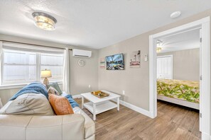 A room with a sofa, a small white table, a wall-mounted air conditioner, a window with curtains, a flat-screen TV, and a door leading to another room. - Walk to Siesta Key Beach & Village | Pool | Dock | Game Room! (Siesta Key)