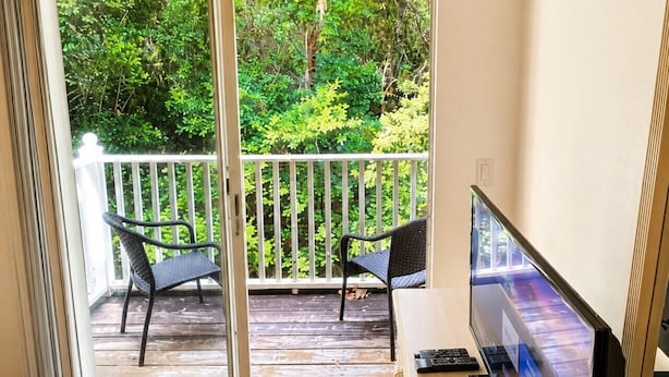Une chambre d’hôtel avec un lit, un bureau avec une télévision et un balcon équipé de chaises, offrant une vue sur la verdure.