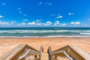 Ein hölzerner Steg führt zu einem Strand mit blauem Himmel und Meereswellen.