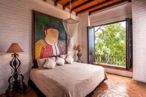 Ein Schlafzimmer mit einem großen Bett, einem Gemälde an der Wand, zwei Nachttischlampen und Blick durch ein Fenster.