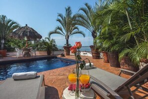 Ein Poolbereich mit Liegestühlen, einem Tisch mit Rose und Getränken sowie einer strohgedeckten Struktur.