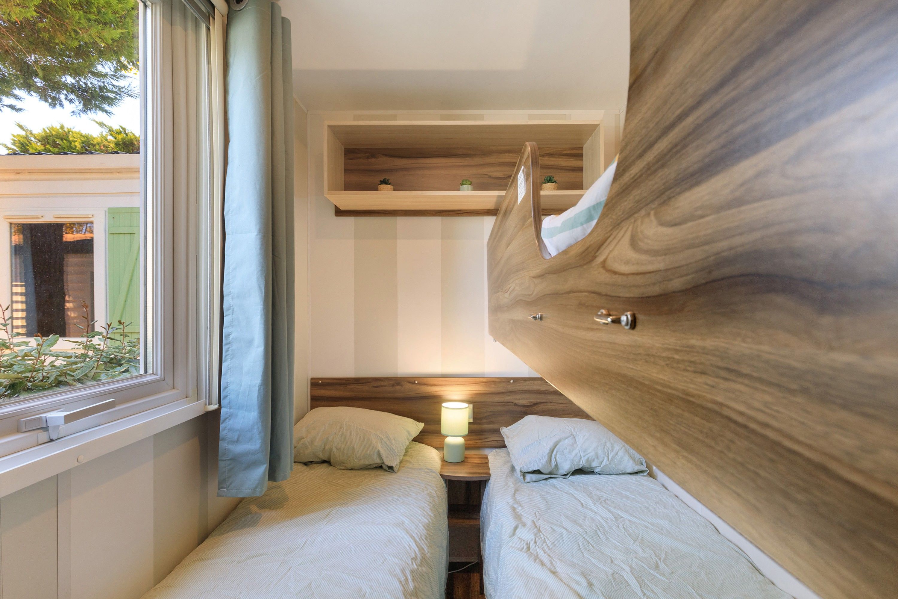 Une chambre avec deux lits, une fenêtre avec des rideaux, une table de chevet avec une lampe et une tête de lit en bois.