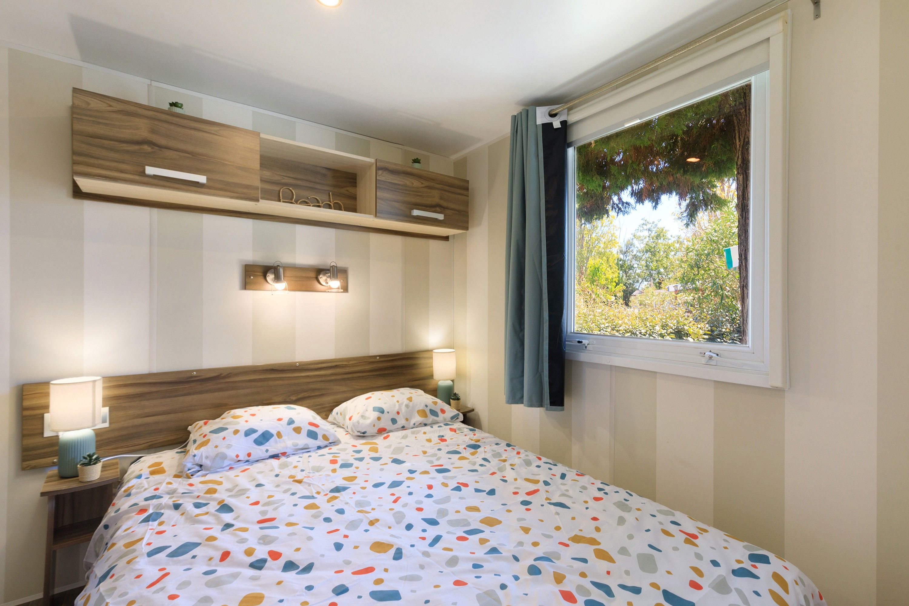 Une chambre à coucher comprenant un lit, des lampes de chevet, une fenêtre avec des rideaux et des rangements intégrés en bois.