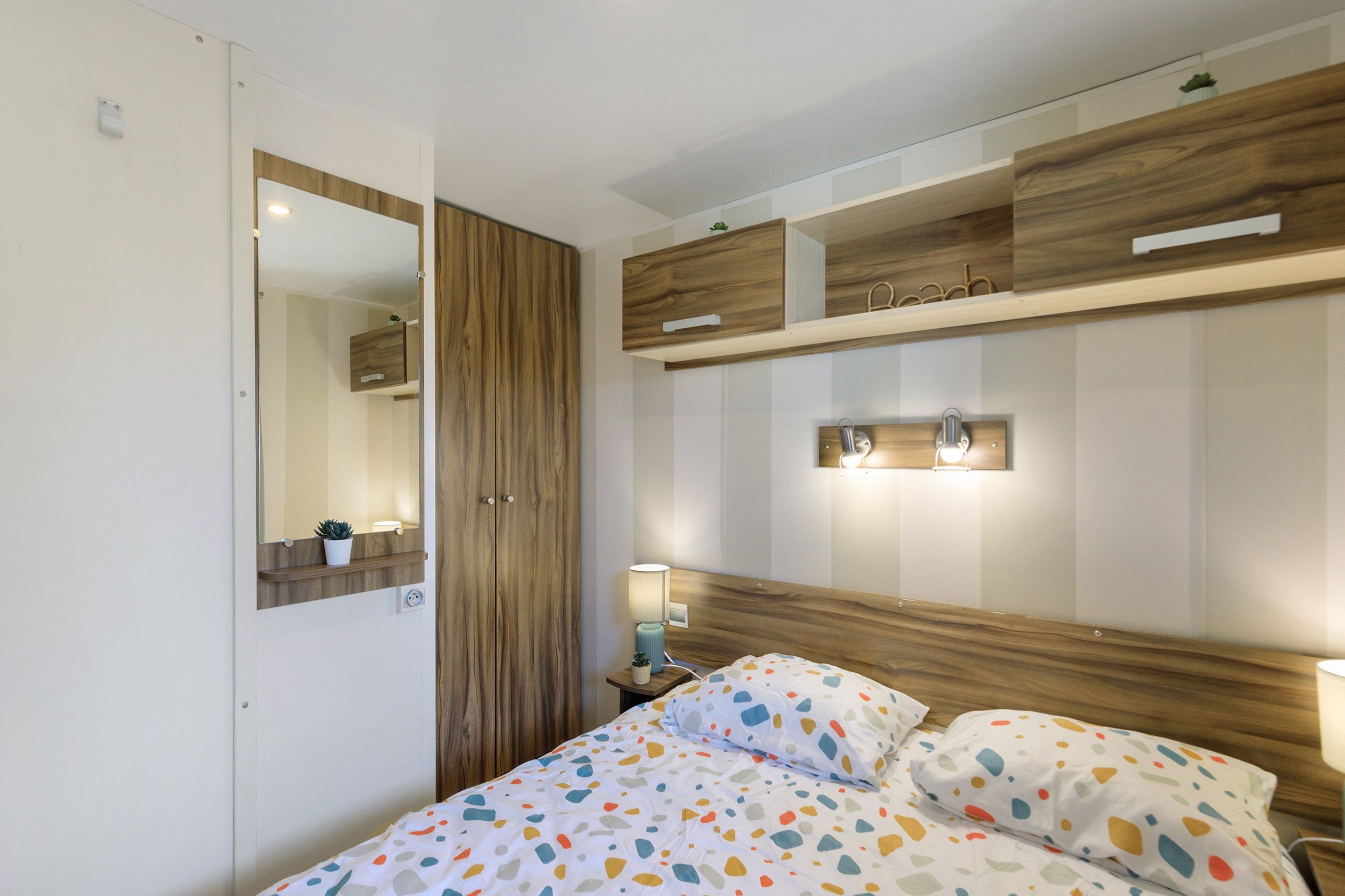 Une chambre à coucher comprenant un lit, une armoire en bois, un miroir, une table de chevet avec une lampe, et une étagère murale agrémentée d’objets décoratifs.