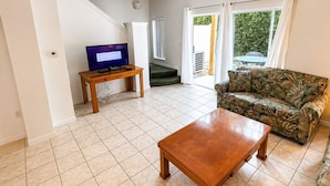 Una sala de estar con una mesa de centro de madera, un sofá, un televisor sobre un mueble bajo y una escalera que lleva a un espacio al aire libre.