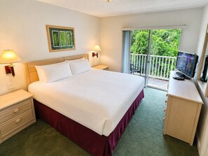A hotel room with a bed, bedside lamps, a desk with a television, and a balcony with a view of greenery. - Your Next Adventure 3 (Kissimmee)