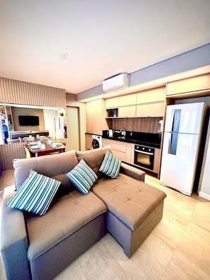 Uma sala de estar moderna com um sofá seccional, uma TV de tela plana, uma cozinha com geladeira, forno e micro-ondas.