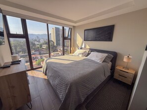 Un dormitorio con un ventanal grande, una cama con colcha gris, una mesita de noche, una lámpara y una obra de arte colgada en la pared.