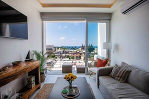 A modern living room with a sofa, TV, wooden console, and a view of the city and sea through large glass doors. - Vrede Penthouse | Ocean Views • Rooftop Pool (Puerto Vallarta)