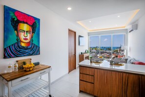 A modern kitchen with a large window, a wooden island, and a painting on the wall. - Vrede Penthouse | Ocean Views • Rooftop Pool (Puerto Vallarta)