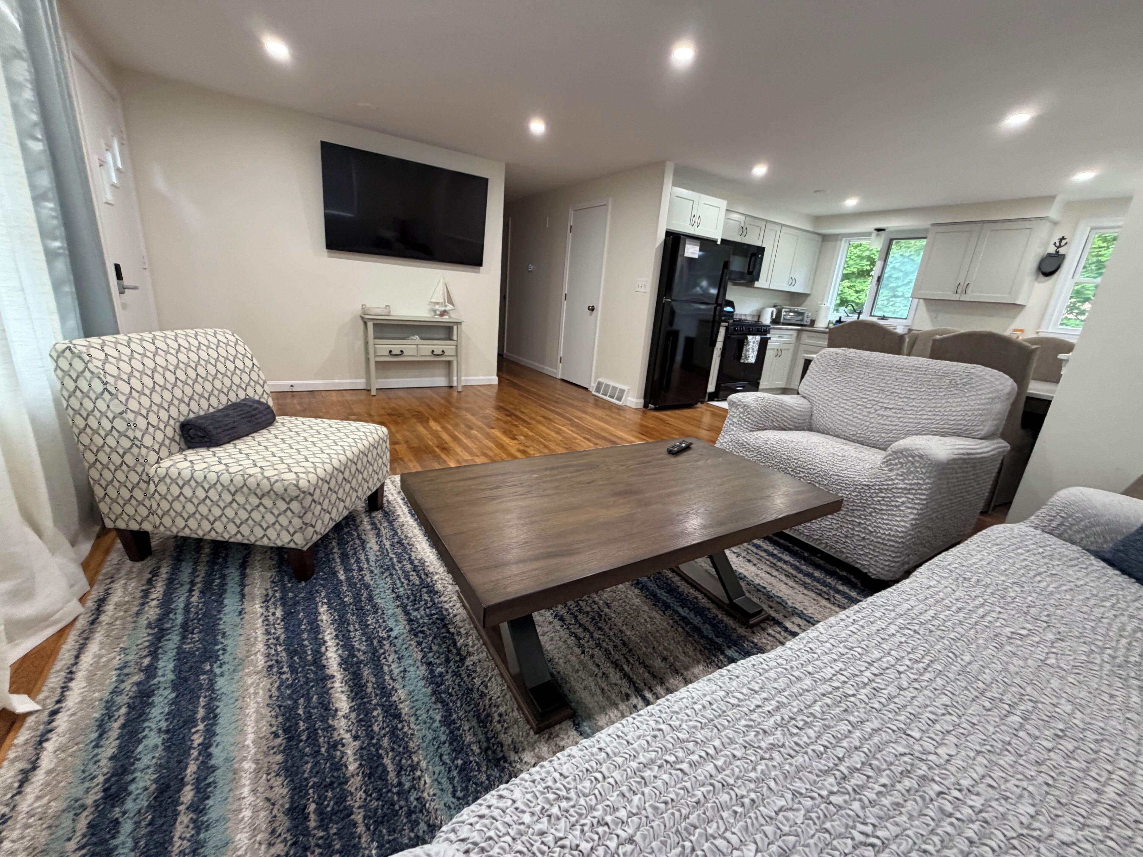 Ein modernes Wohnzimmer mit Sofa, Sessel und Couchtisch, im Hintergrund eine Küche und ein Flachbildfernseher an der Wand.